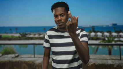 Man in striped shirt shows middle finger on street terrace overlooking sea coast under blue sky;...