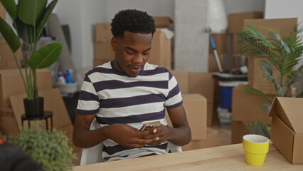 Man taps smartphone while seated at wooden table in new home living room with cardboard boxes;...