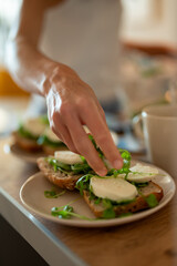 Close-up of a hand preparing healthy open sandwiches with fresh mozzarella and arugula on whole-grain bread, creating a light nutritious breakfast in a cozy home kitchen