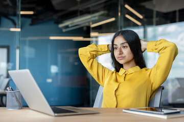A thoughtful and serious young Indian woman is sitting in the office at her desk, her hands behind her head, and looking at the laptop screen