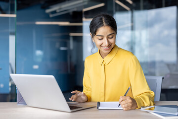 Smiling young Indian businesswoman working in the office at a laptop and taking notes in a notebook