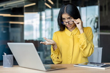 A stressed young Indian businesswoman is looking at her laptop screen in the office, lowering her glasses with her hand and spreading her arms