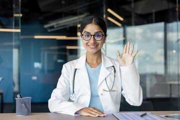 A young Indian female doctor sits in an office at a table facing the camera, greeting and conducting an online consultation
