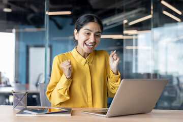 A young Indian woman sits at a desk in the office, looks at her laptop and rejoices in her success