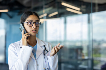 Close-up photo of a young Indian female doctor standing in a white coat in a clinic and talking on the phone