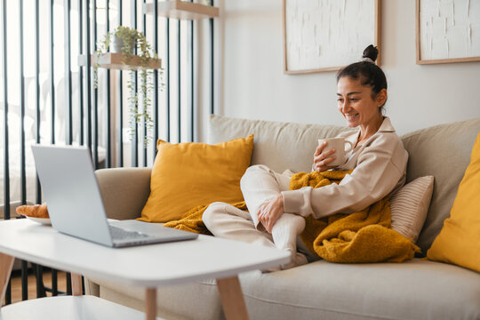 Woman relaxing on a cozy sofa wrapped in a warm mustard blanket, looking thoughtfully at her laptop on the coffee table with a cup and croissants creating a calm and homely atmosphere. - Powered by Adobe