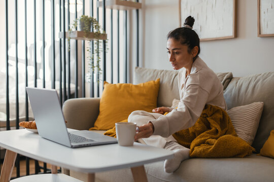 Woman relaxing on a cozy sofa wrapped in a warm mustard blanket, looking thoughtfully at her laptop on the coffee table with a cup and croissants creating a calm and homely atmosphere. - Powered by Adobe