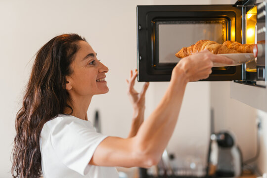 Smiling woman placing a plate with fresh croissants into a microwave oven in a bright modern kitchen, enjoying a relaxed morning moment while preparing a warm breakfast at home.