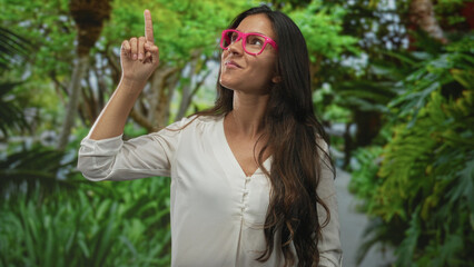 Obraz premium Woman wearing pink glasses and white blouse gives thumbs up in a forest walkway, smiling at camera; confidence optimism.