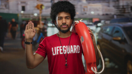 Lifeguard man making oath gesture on a city street at night surrounded by urban lights, wearing red shirt and holding safety buoy, symbolizes promise and readiness for duty outdoors.