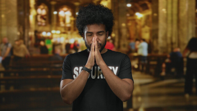 Man wearing security uniform with eyes closed in prayer inside church with blurred interior background offering a serene and reflective atmosphere.