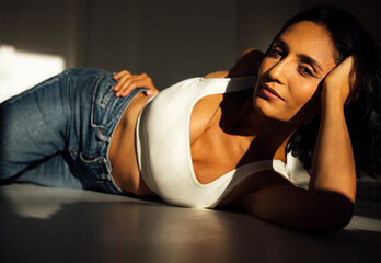 Beautiful woman with dark hair, wearing a white top and blue jeans, poses gracefully on a surface,...