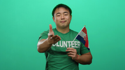 Young man holds a croatian flag against a green background while wearing a volunteer shirt.