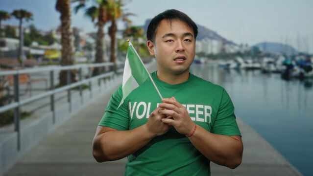 Young man holding nigerian flag on seaside promenade, expressing nervousness, wearing volunteer shirt outdoors near beach.