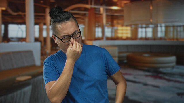Man pinches nose with fingers in restaurant building wearing blue shirt and glasses; disgust smell reaction.