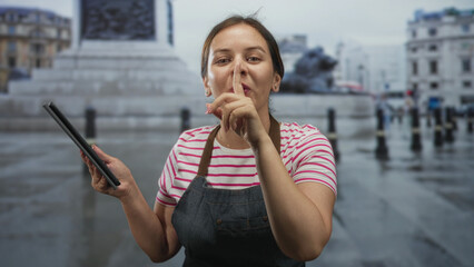 Woman in apron taps tablet and brings finger to lips for silence on street near monument; quiet concentration service.