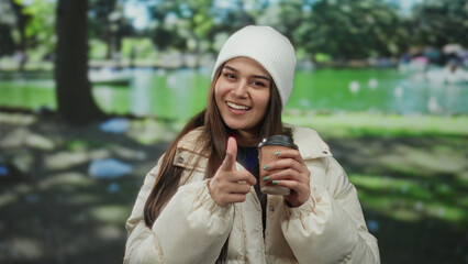 Young woman in a park wearing a white coat and beanie, holding a coffee cup, smiling and pointing...