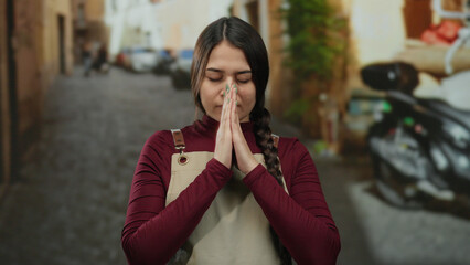 Waitress praying with eyes closed in an outdoor street setting wearing apron peaceful thoughtful serene hispanic woman solitude urban