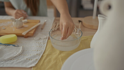 Girl sculpts wet clay on wooden table with hands in art studio, forming small pot beside pottery tools; creative passion.