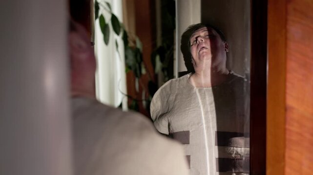 A dim room. A man stares blankly into his own reflection. He is wearing a white straitjacket with long, tied sleeves. The contrast between the motionless body and the inner chaos.