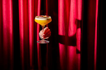 A vibrant yellow cocktail in a coupe glass held by a hand against a luxurious, deep red velvet...