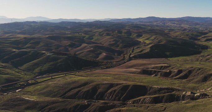 Aerial view of the undulating Craco hills, displaying a tapestry of green and brown, dissected by a long highway, Craco, Basilicata, Italy.