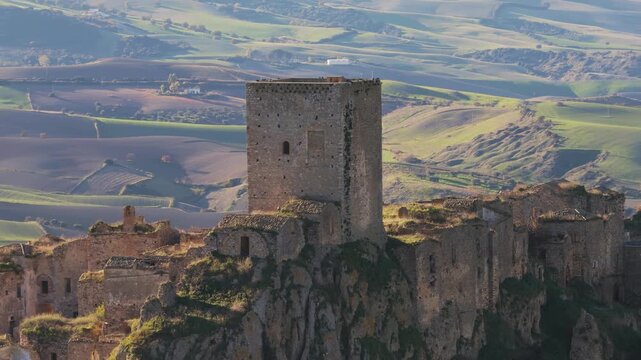 Aerial view of the weathered, ancient stone buildings of Craco old town clinging to a steep hillside with rolling hills in the background, Craco, Basilicata, Italy.