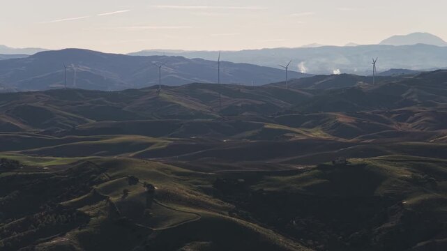 Aerial view of rolling hills dotted with wind turbines creating a stark contrast against the distant mountains, Craco, Basilicata, Italy.