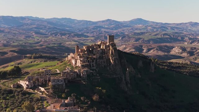 Aerial view of the ancient hilltop village of Craco, its stone buildings contrasting against the green hills and distant mountains, Craco, Basilicata, Italy.