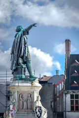 Fototapeta premium Jacob van Artevelde’s bronze statue rises above historic Ghent rooftops, his outstretched arm framed by clouds and architectural details that highlight the city’s medieval heritage.