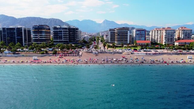 Drone pulling back over a crowded beach and turquoise sea, revealing modern coastal buildings and a long avenue leading toward majestic mountain ranges in Antalya.