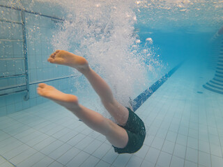 Underwater shot of a young child's legs and feet splashing into a brightly lit indoor swimming pool, creating a burst of bubbles.