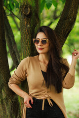 Stylish woman leaning against a tree wearing sunglasses and a beige cardigan in a green park