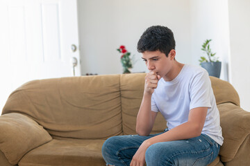 Fototapeta premium A young man sits on a couch, covering his mouth with his hand. He is in a state of distress or discomfort