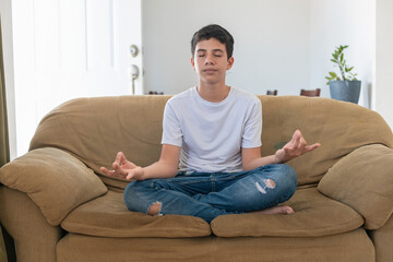 Fototapeta premium A boy is sitting on a couch and practicing yoga. The couch is brown and the boy is wearing a white shirt
