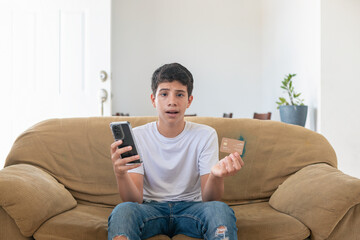 Obraz premium A young man sits on a couch holding a cell phone and a credit card. He looks confused and unsure of what to do with the two items