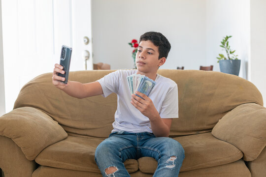 A young boy is sitting on a couch and taking a selfie with his cell phone. He is holding a pile of cash in his hand. Concept of wealth and luxury, as the boy is surrounded by money