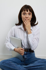 A surprised young woman sits cross legged on the floor, holding a laptop and covering her mouth, conveying shock, curiosity, or unexpected news.