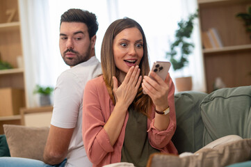 Woman showing surprised reaction looking at smartphone with jealous man