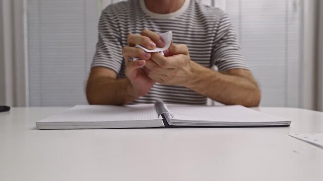 A person tears out a page from a spiral notebook, crumples it into a ball, and discards it on a white desk. This action is repeated, showing frustration or discarding a bad idea.