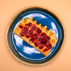Top view of a waffle with strawberry jam on a blue plate, on a warm beige&ndash;orange background.
