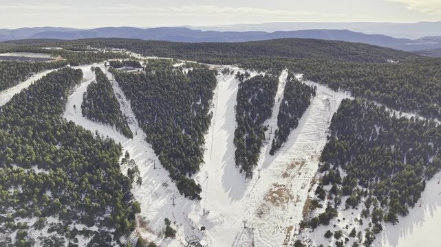 Aerial footage of a drone slowly pulling away from a wide snowy ski slope framed by evergreen pine forest, revealing expansive winter mountains, ski tracks, resort area, serene natural landscape and w