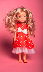Doll with long curly hair wearing a red outfit with white polka dots on a pink background