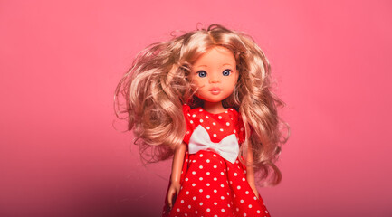 Doll with long curly hair wearing a red outfit with white polka dots on a pink background