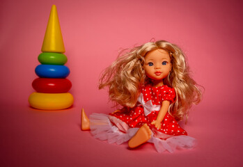 A doll in a red dress with white dots sits on the floor. Nearby is a colorful stack of rings. The setting has pink walls and natural light.