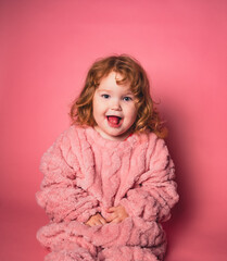 A young child sits on the floor wearing a pink fleece outfit. She smiles happily at the camera with her tongue out, enjoying indoor playtime. The background is pink, adding to the fun. © Anton Dios