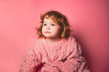 A young child with curly red hair sits against a solid pink background. The child wears a fuzzy pink outfit and shows a thoughtful expression.