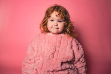 A young child with curly red hair sits against a solid pink background. The child wears a fuzzy pink outfit and shows a thoughtful expression.