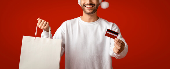 Man wearing a festive hat holds a shopping bag and credit card against a bright red background. He is happily preparing for holiday shopping, showcasing seasonal excitement.