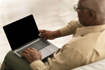 An older man is seated on a couch, focused on his laptop in a warm and inviting living room. The light creates a peaceful atmosphere for his activity.